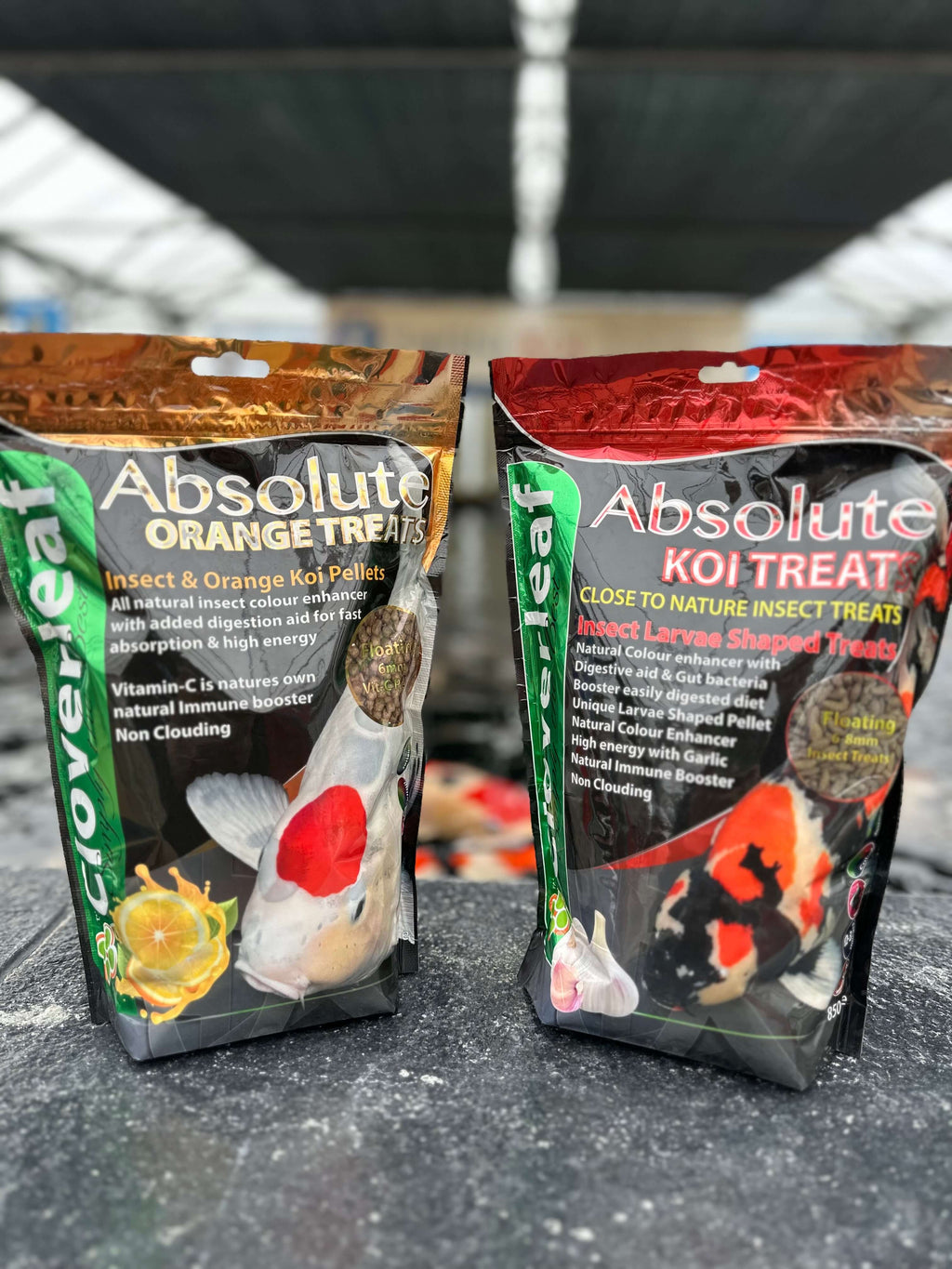 Cloverleaf Koi Treats Insect Larvae Garlic packaging with orange and koi treat varieties in a garden center environment.